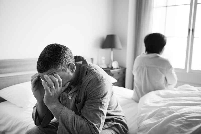 Man with his head in his hands sitting on one side of the bed, while a woman sits on the other side facing the opposite direction.